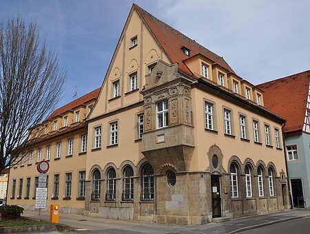Gebäude B des Landratsamts Weißenburg-Gunzenhausen Historisches Gebäude mit gelber Fassade, roten Dachziegeln und Erker an Straßenecke bei bewölktem Himmel.